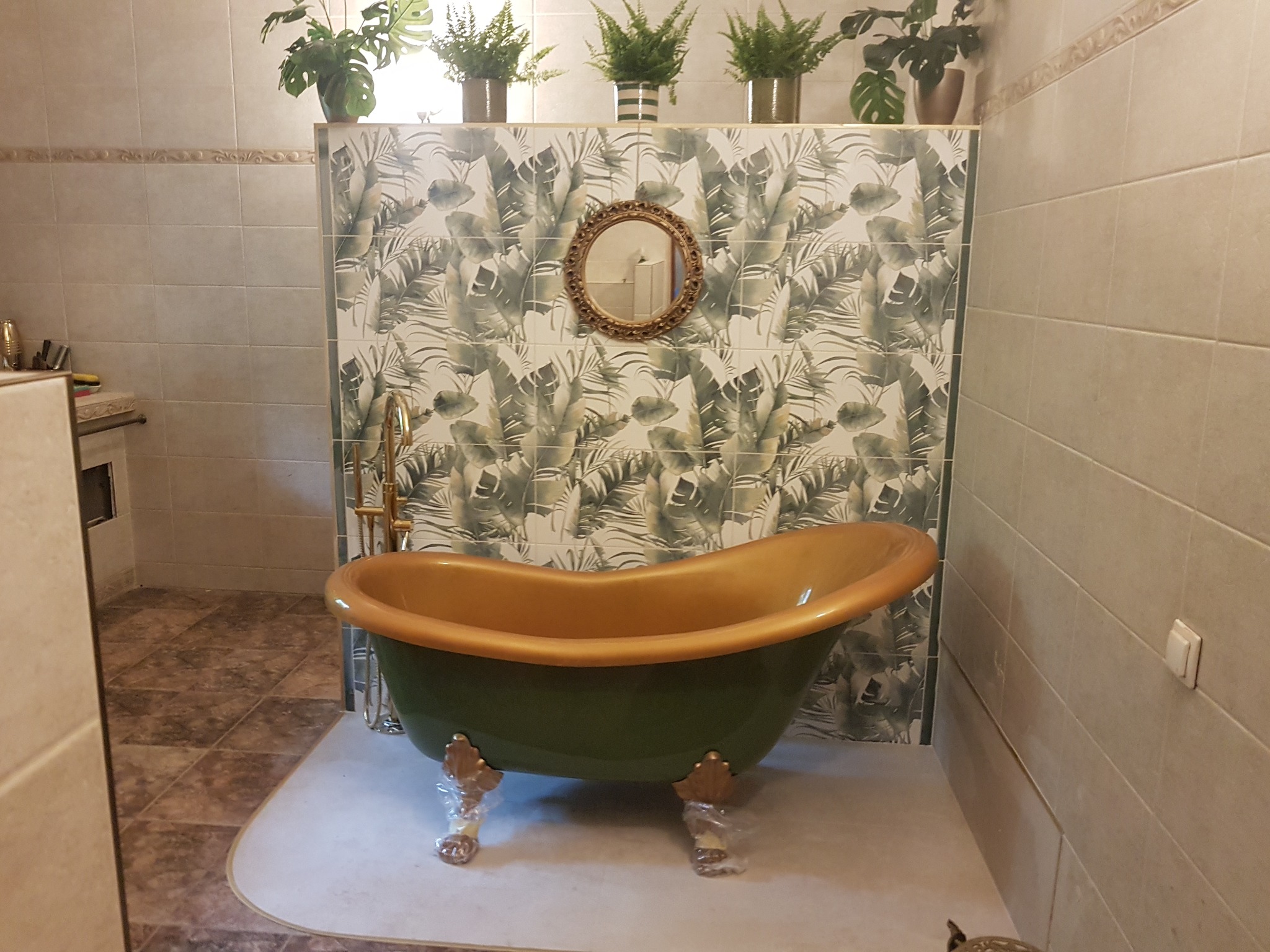 Oliwkowa wanna wolnostojąca z pozłacanym wnętrzem i nóżkami w folii ochronnej, na tle ściany w tropikalne liście i okrągłego lustra w złotej ramie, w jasnej łazience.