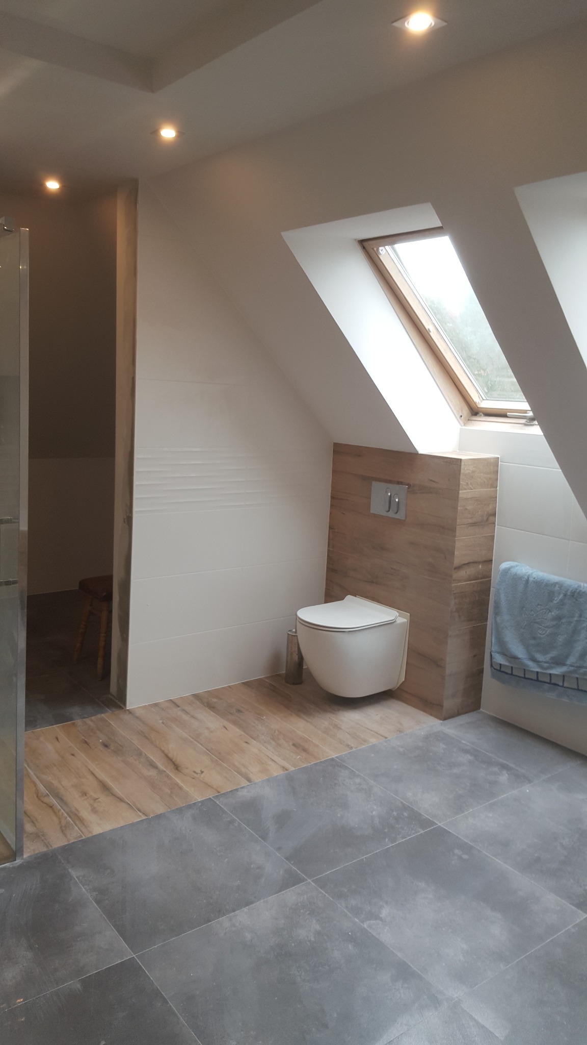 Nowoczesna łazienka na poddaszu z oknem dachowym, drewnopodobnymi płytkami na ścianie za podwieszaną toaletą i przejściem do strefy prysznica, gdzie widoczny jest drewniany stołek.