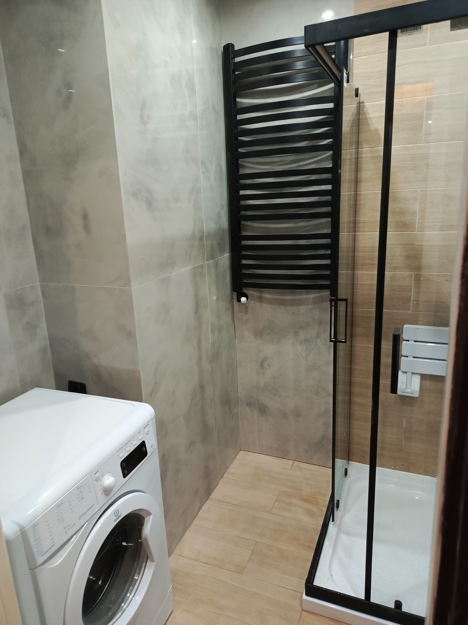 Wykończona łazienka z marmurowymi ścianami, białą pralką Indesit, czarnym grzejnikiem drabinkowym i kabiną prysznicową z czarnymi profilami oraz siedziskiem.