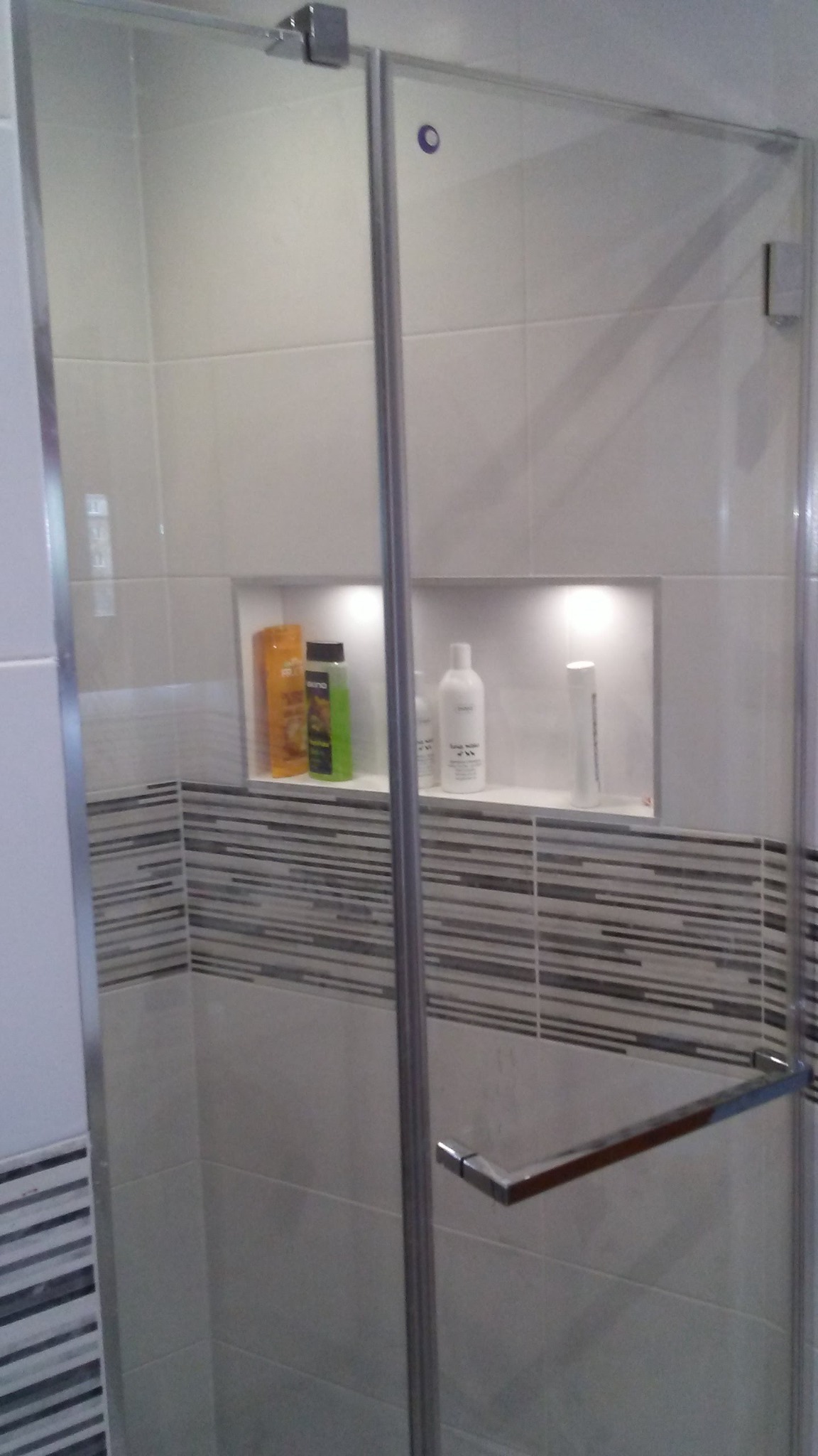 Szklana kabina prysznicowa z chromowanymi okuciami, widoczne półki w ścianie z kosmetykami, dekoracyjne pasy płytek.