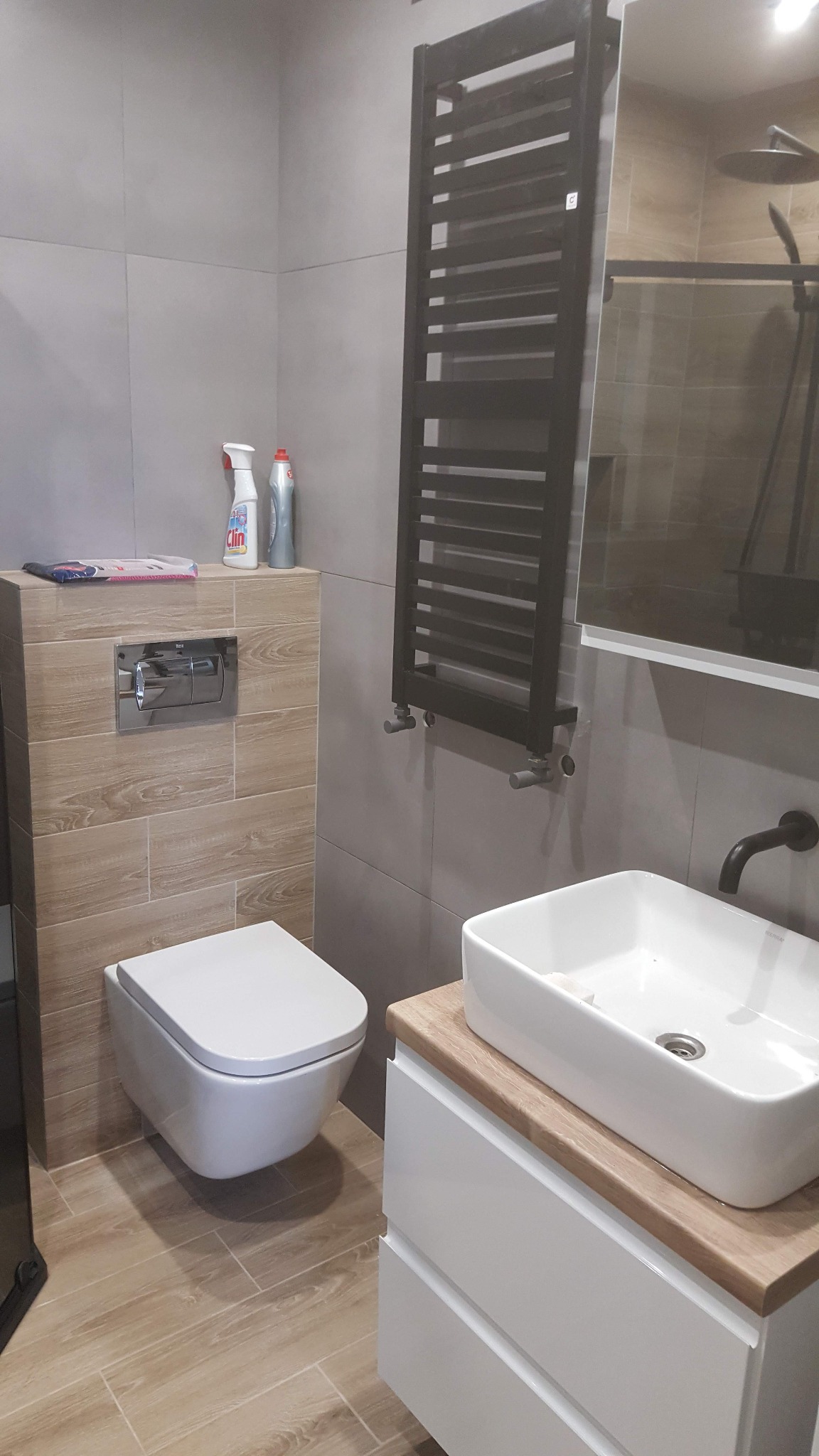 Nowoczesna łazienka z szarymi płytkami, drewnopodobnymi akcentami, wiszącą toaletą, grzejnikiem drabinkowym i umywalką nablatową na szafce z drewnianym blatem.