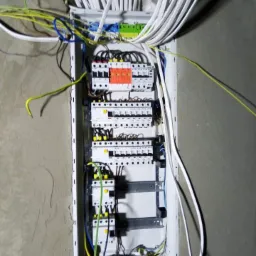 Rozdzielnica elektryczna w trakcie montażu w surowym, szarym pomieszczeniu, widoczne wystające przewody w różnych kolorach i grubościach, oraz fragment wybitej ściany nad obudową.