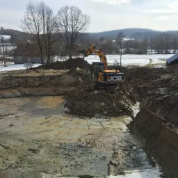 Żółta koparka JCB na placu budowy w trakcie prac ziemnych, widoczne wykopane fundamenty, w tle krajobraz wiejski z ośnieżonymi polami i domami.