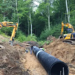 Dwa żółte i czarne koparki Liebherr i JCB pracują przy układaniu długiej, czarnej rury w wykopie na tle zielonego lasu. Dwóch pracowników nadzoruje proces.