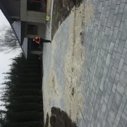 Układanie szarej kostki brukowej na posesji, widoczny fragment przygotowanego podłoża z piaskiem, pracownik w pomarańczowej kamizelce, w tle dom i tuje.