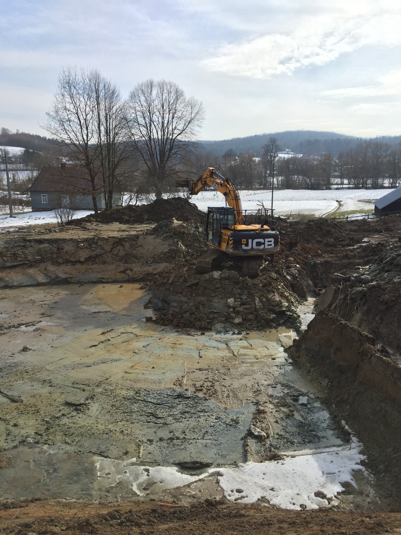 Żółta koparka JCB na placu budowy w trakcie prac ziemnych, widoczne wykopane fundamenty, w tle krajobraz wiejski z ośnieżonymi polami i domami.