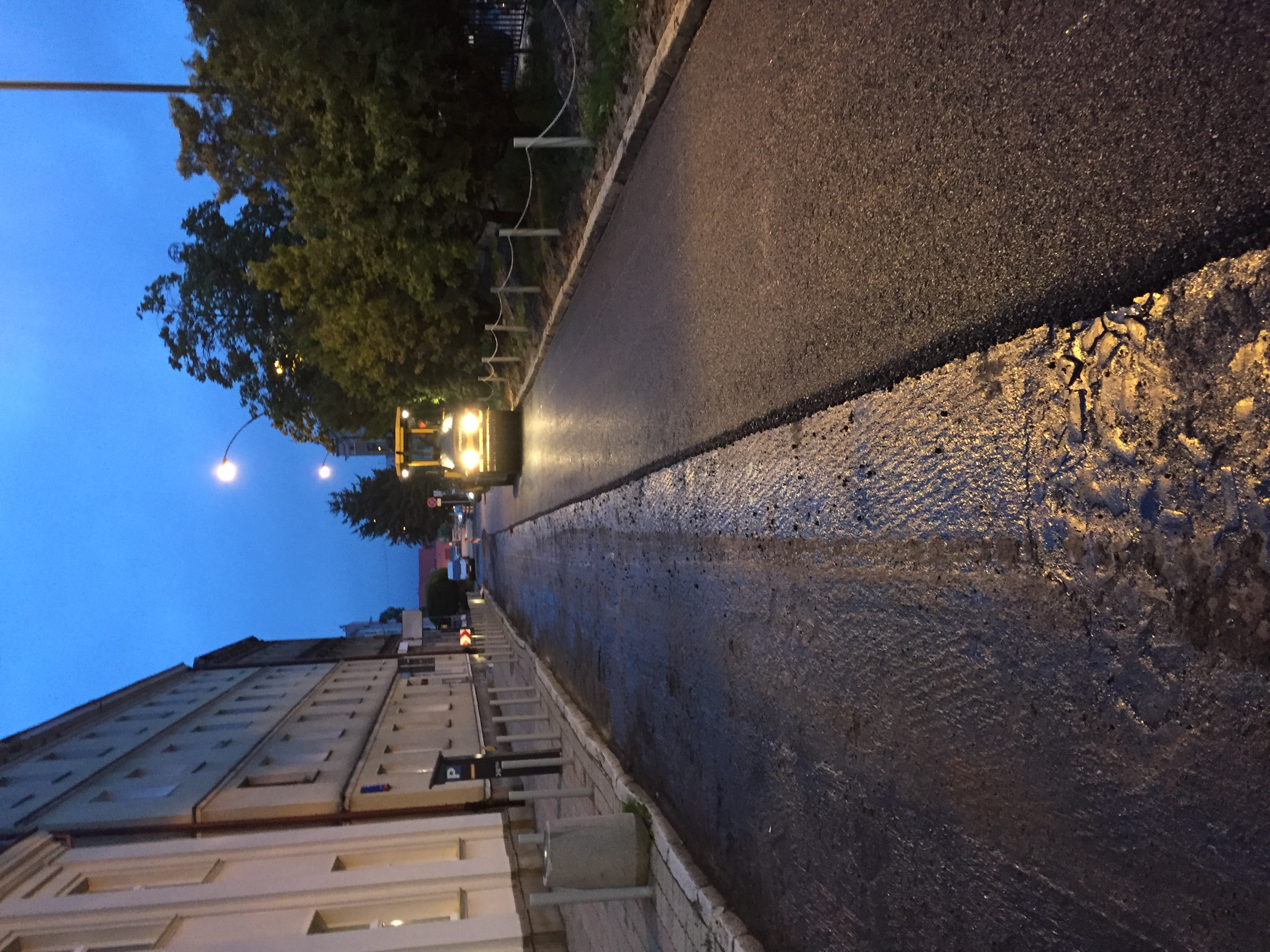 Walec drogowy z włączonymi światłami ubija świeżo wylany asfalt na ulicy miejskiej o zmierzchu.