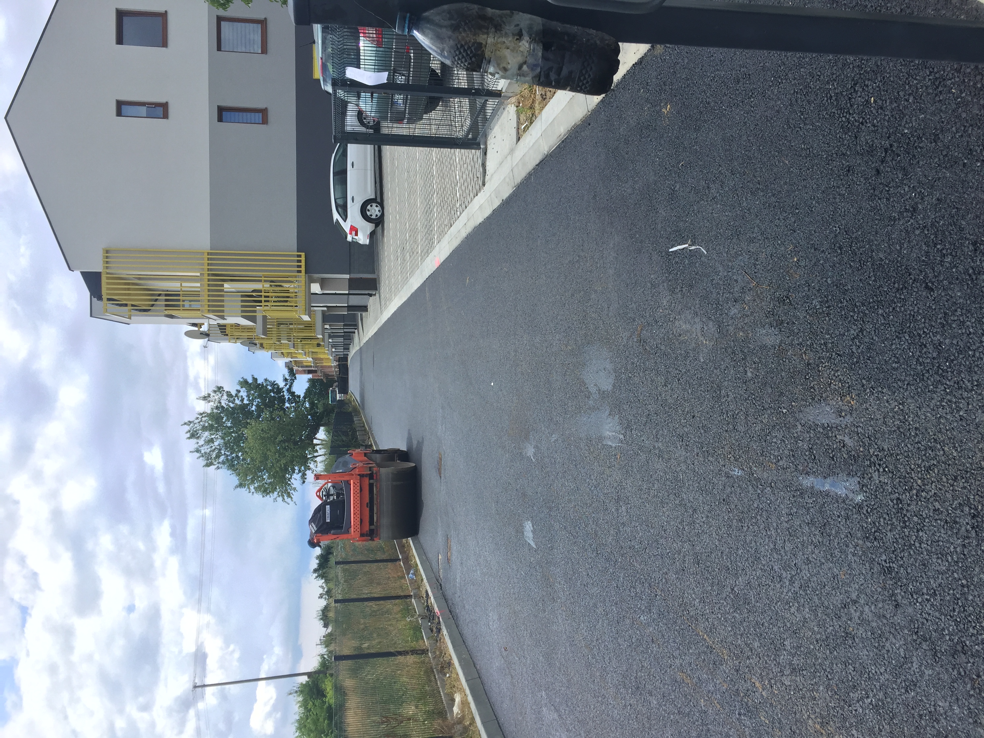 Walec drogowy podczas zagęszczania świeżo wylanego asfaltu na drodze osiedlowej, z widocznymi budynkami mieszkalnymi w tle.