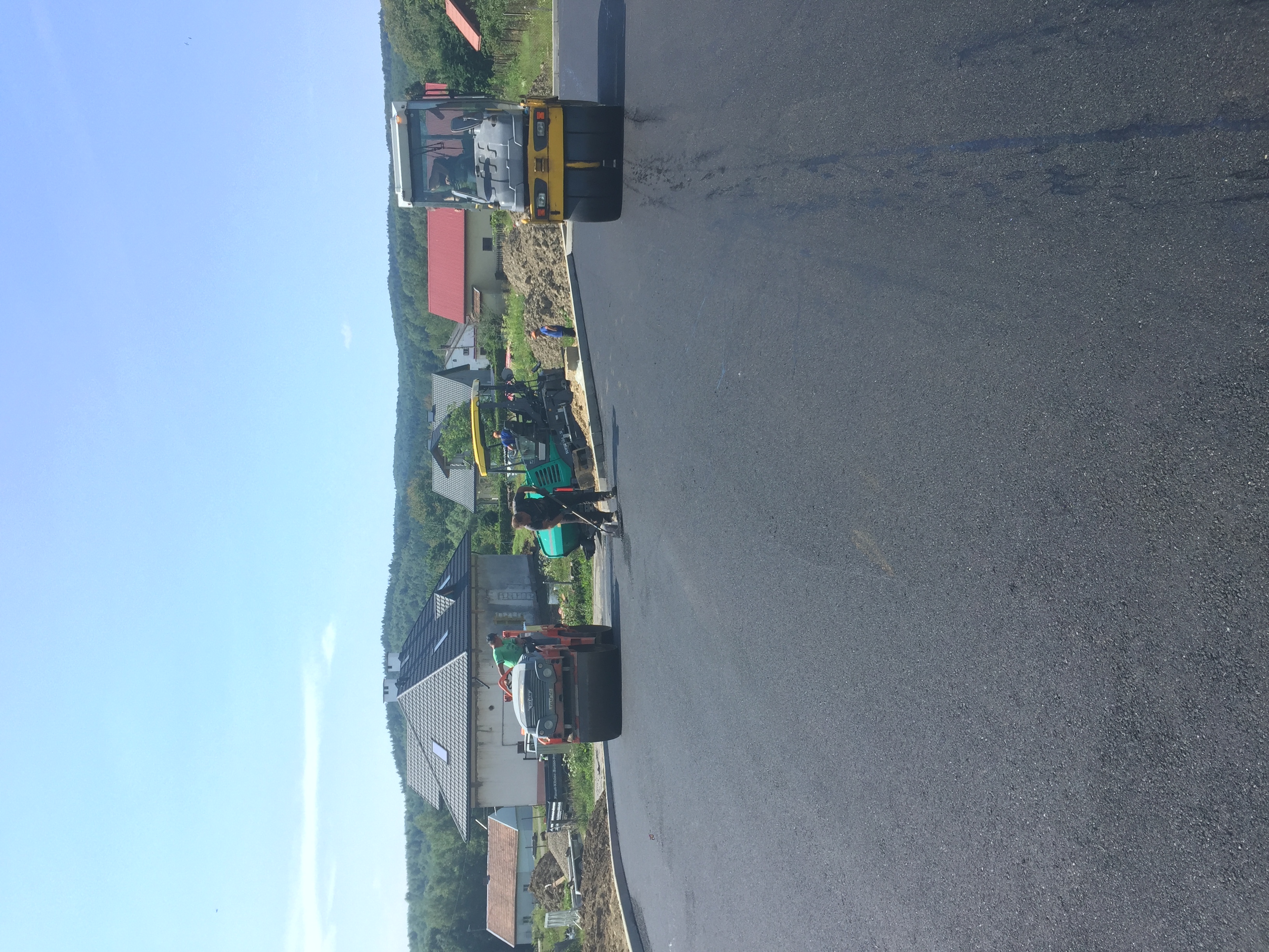 Walec drogowy ugniata świeżo wylany asfalt na nowej drodze, w tle zabudowa jednorodzinna.