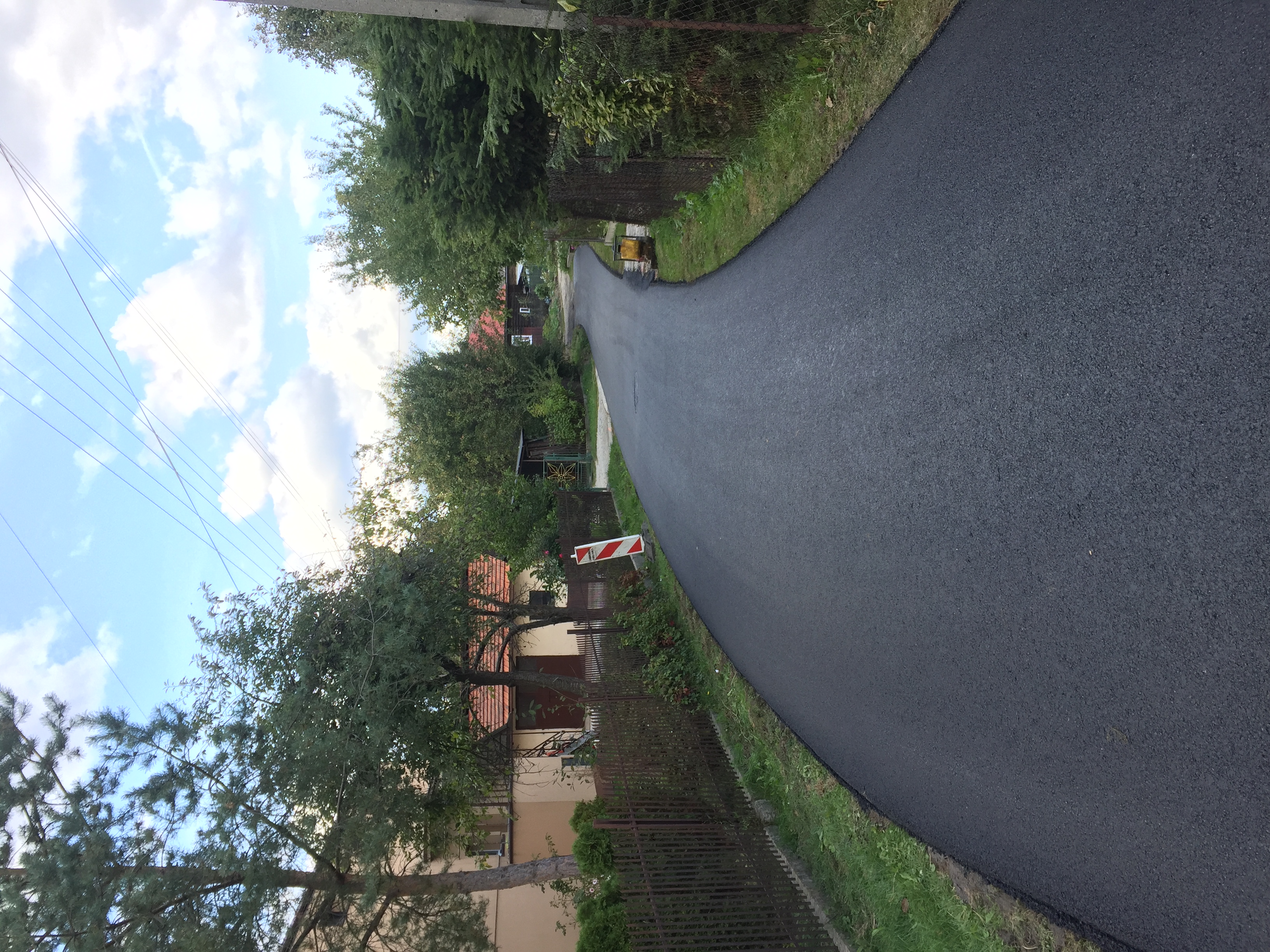 Nowa, gładka nawierzchnia asfaltowa drogi osiedlowej, widok z poziomu gruntu, z widocznym oznakowaniem tymczasowym i zielenią po bokach.