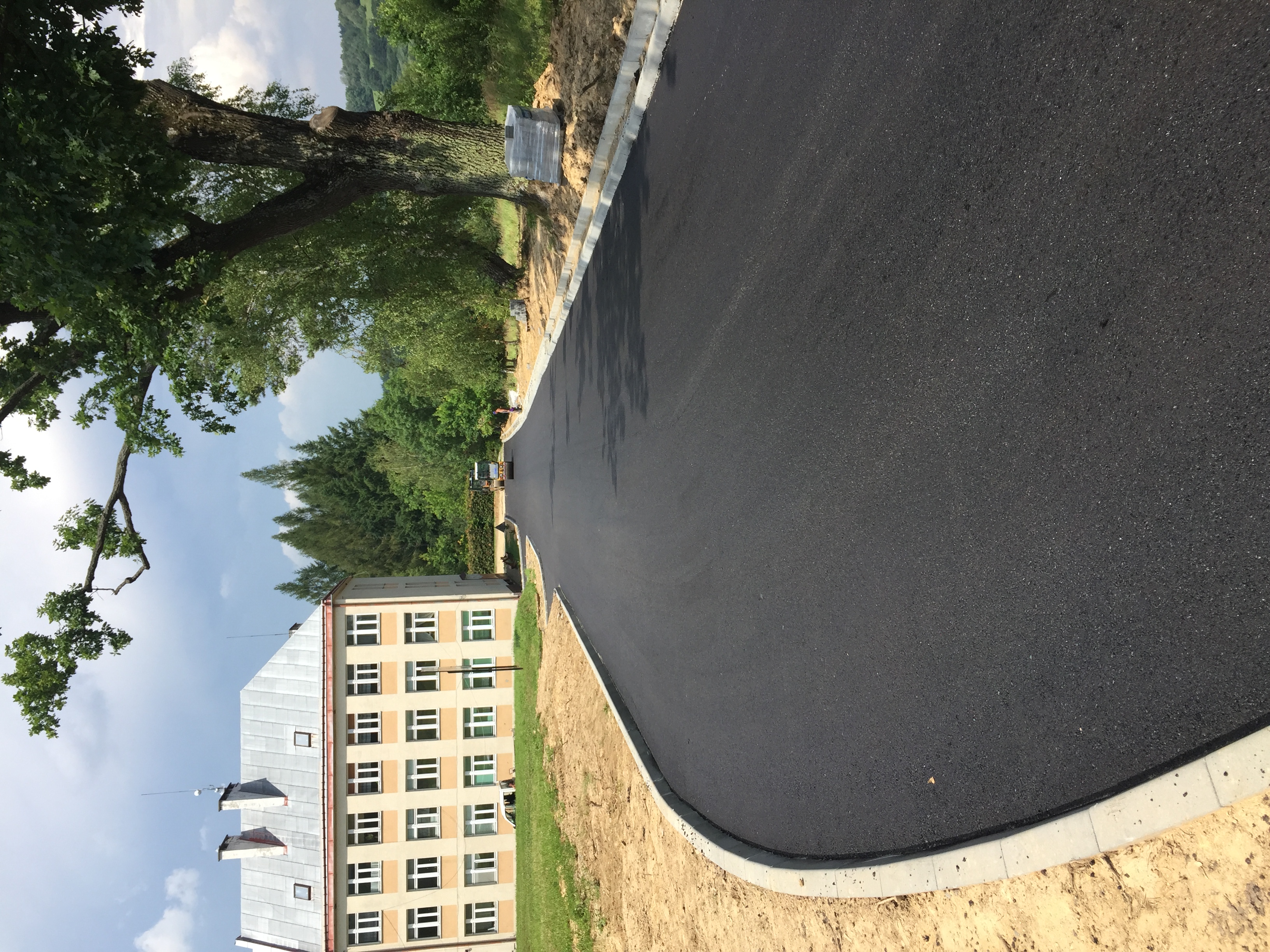 Nowo wyasfaltowana droga z betonowymi obrzeżami, obok drzewo i budynek mieszkalny w tle.