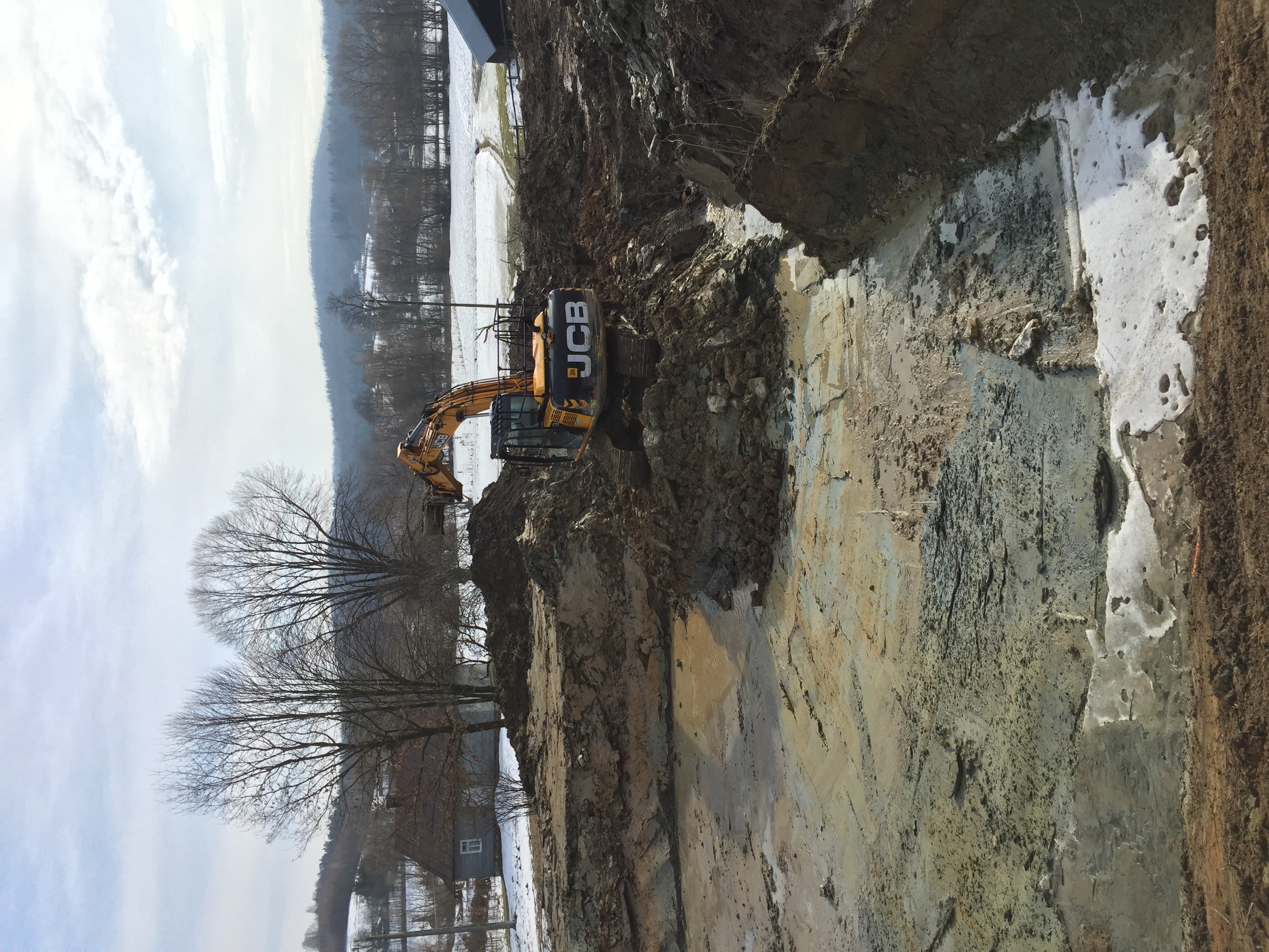 Żółta koparka JCB w trakcie prac ziemnych, widoczny wykop w gruncie z resztkami śniegu i błota, w tle wiejski krajobraz z drzewami i zabudowaniami.