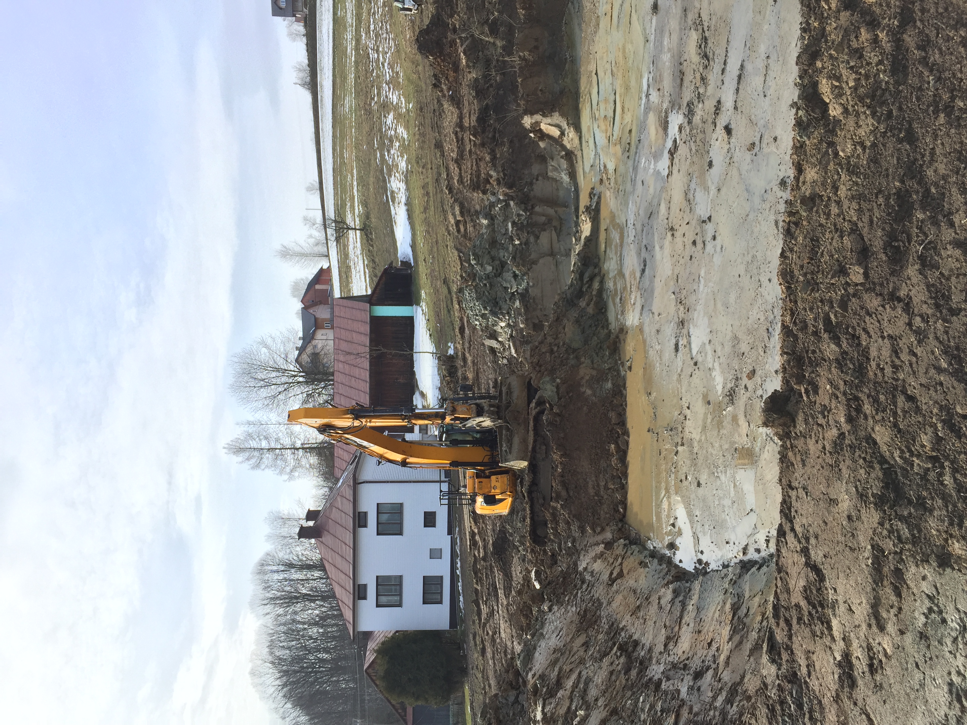 Żółta koparka wykonuje wykop w błotnistym terenie, w tle biały dom z czerwonym dachem i zaśnieżone pole.