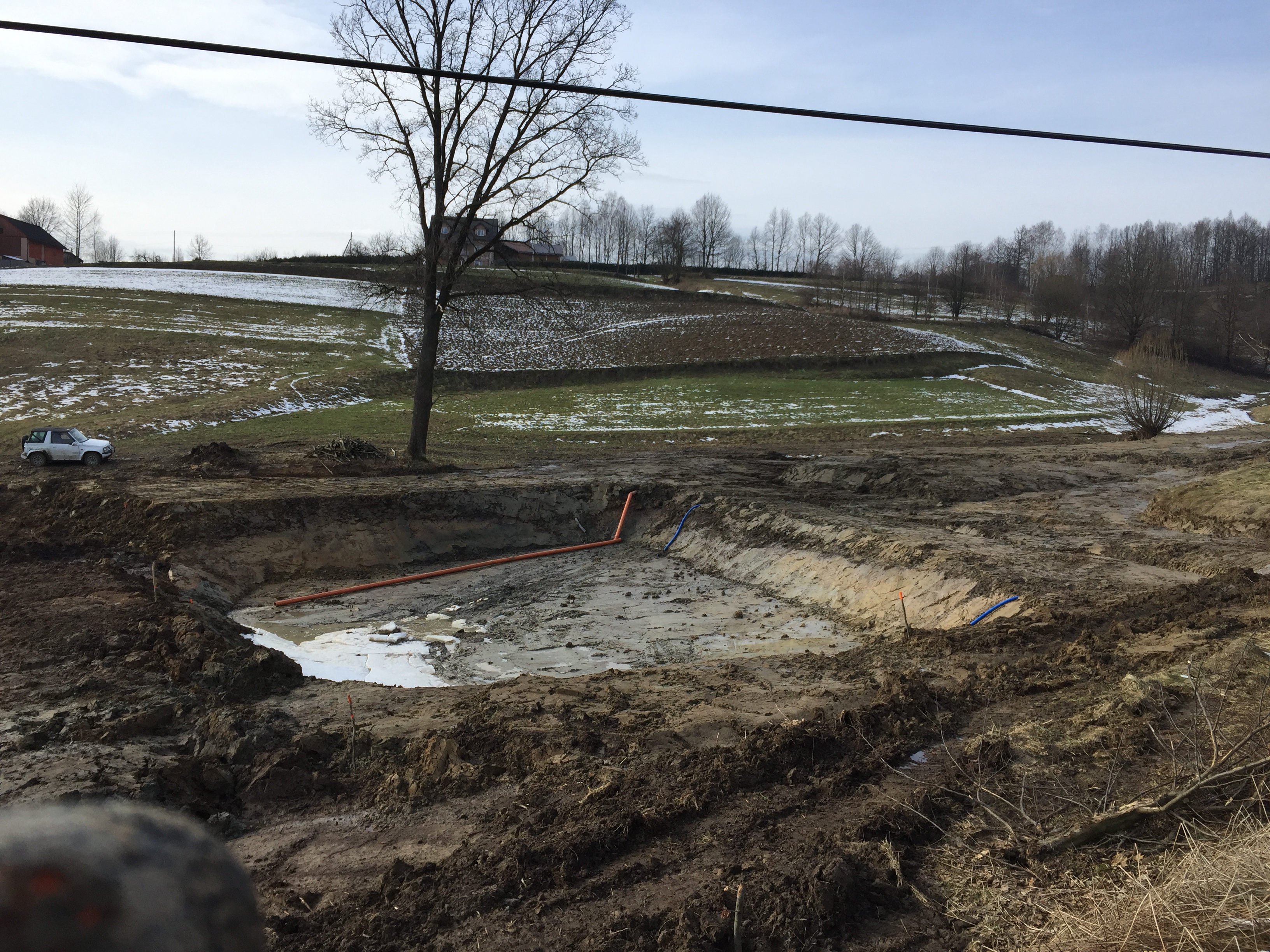 Wykonany wykop pod zbiornik na wodę z widocznymi rurami w ziemi, w tle zimowy krajobraz z resztkami śniegu i samochodem