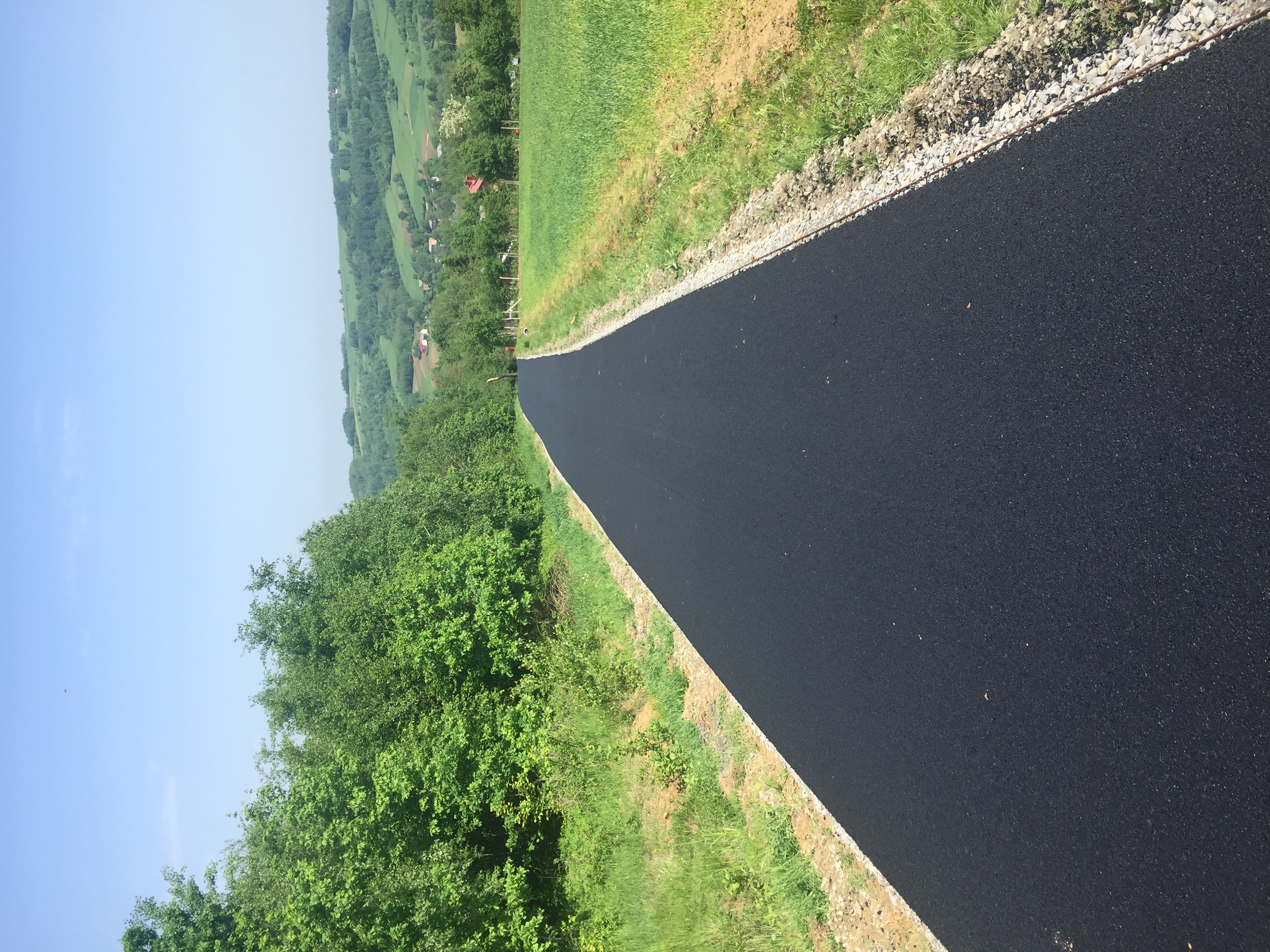 Świeżo położona, czarna nawierzchnia asfaltowa drogi polnej, z widocznym poboczem z kamieni i zielenią łąk po bokach, w oddali zalesione wzgórza i błękitne niebo.