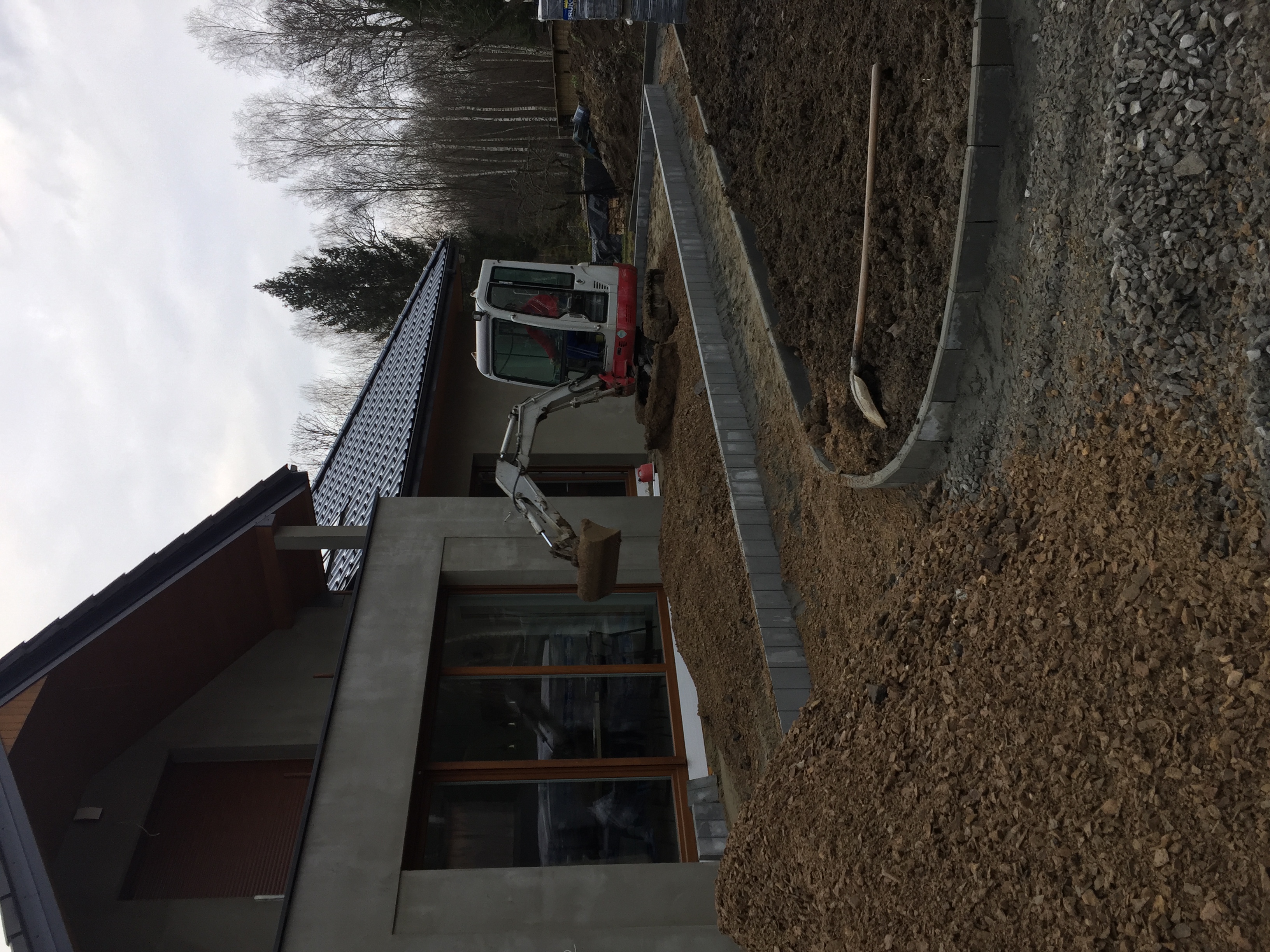 Mini koparka wykonuje wykop wzdłuż fundamentów domu, przygotowując teren pod chodnik z kostki brukowej, widoczny świeżo ułożony obrzeżnik i podsypka.