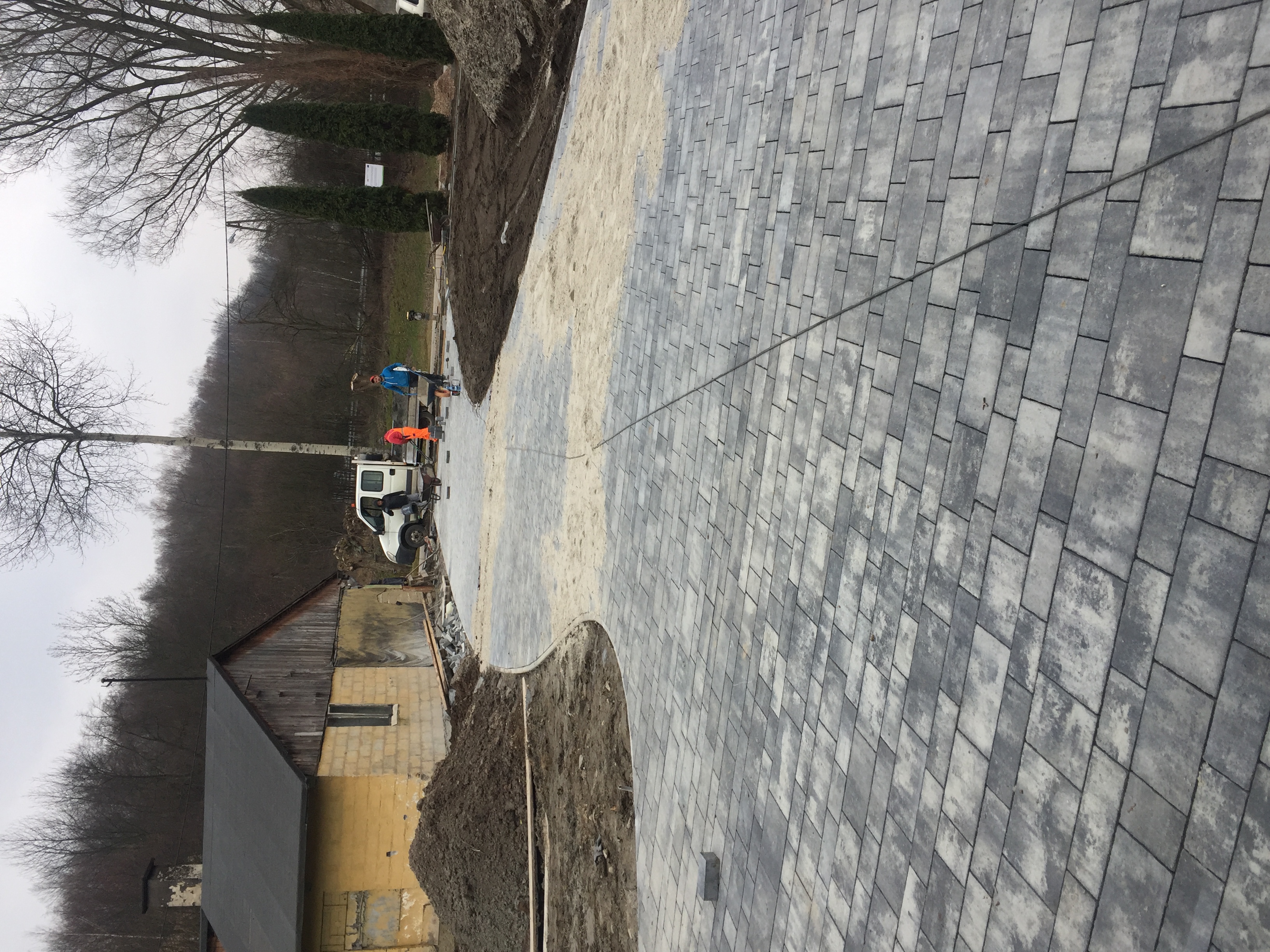 Układanie szarej kostki brukowej w Golcowej, widoczna część drogi z ułożoną kostką, pracownicy i samochód w tle.