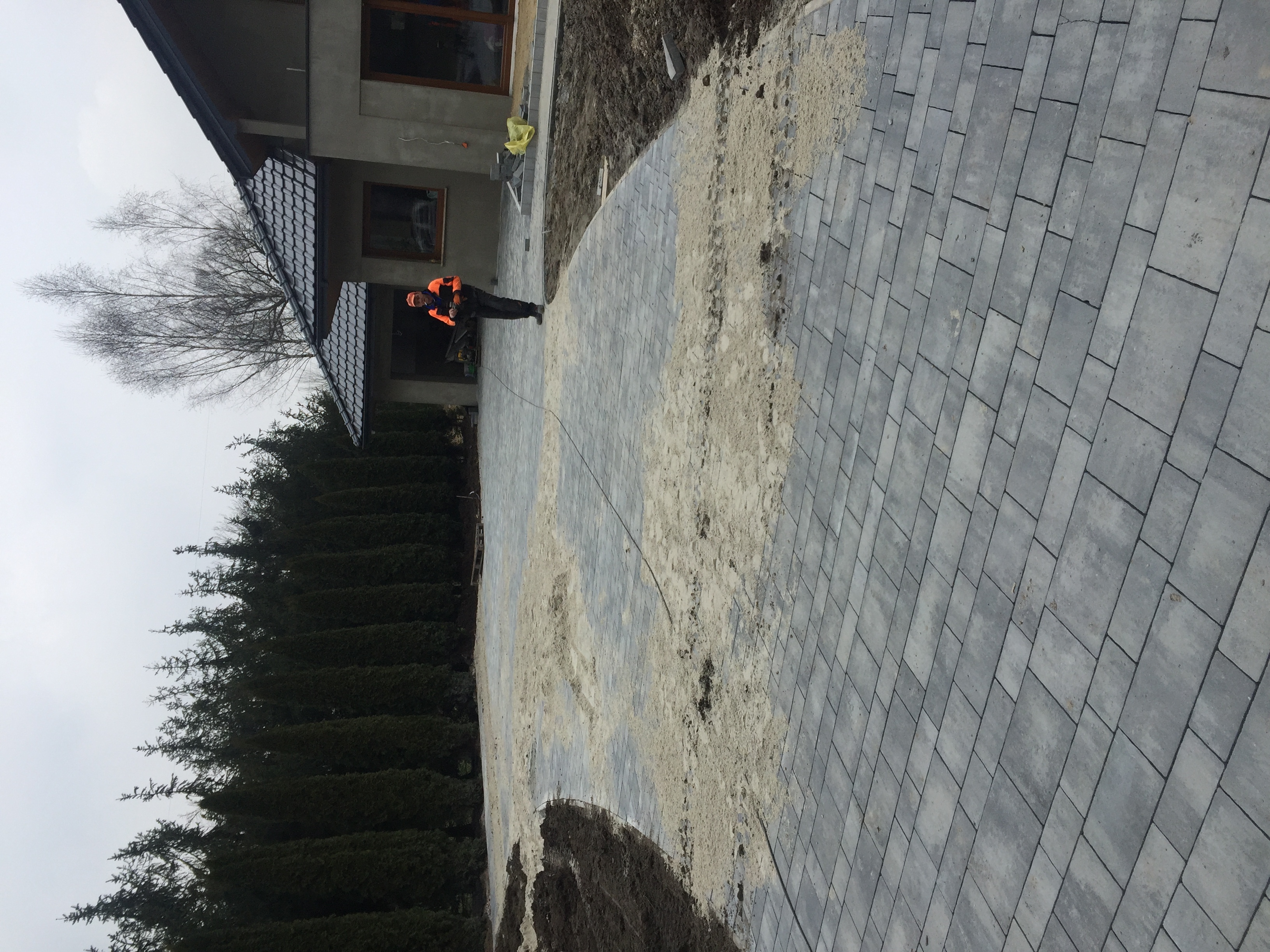 Układanie szarej kostki brukowej na posesji, widoczny fragment przygotowanego podłoża z piaskiem, pracownik w pomarańczowej kamizelce, w tle dom i tuje.