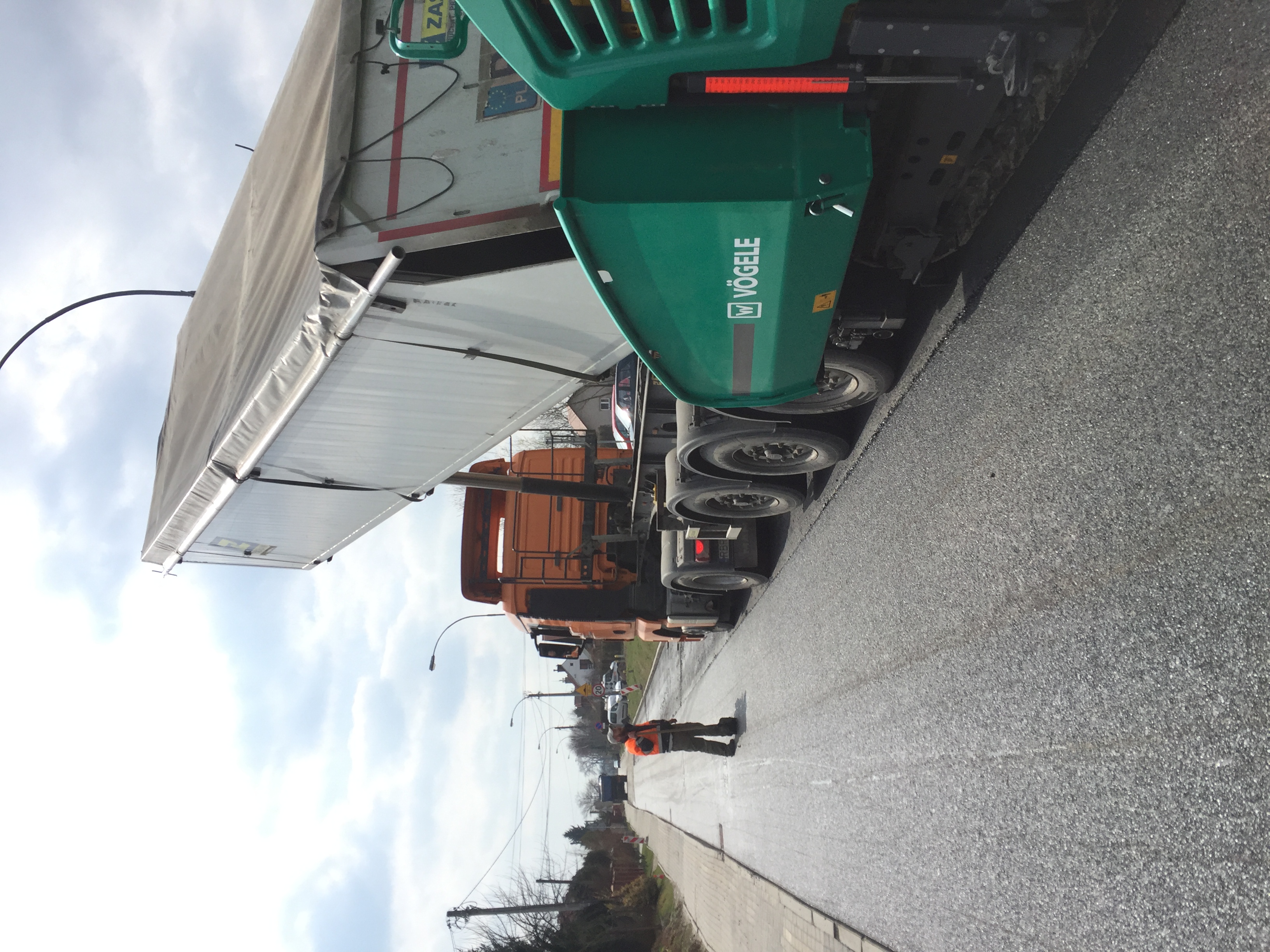 Układanie asfaltu na drodze za pomocą rozściełacza Vögele i wywrotki, pracownik w kamizelce odblaskowej nadzoruje proces.