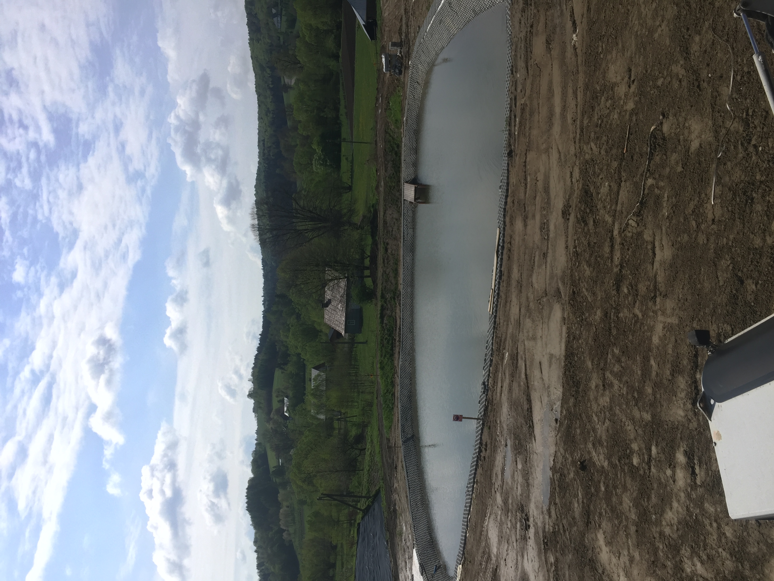 Nowo wykopany, częściowo wypełniony wodą staw z zabezpieczonymi brzegami, widok na pagórkowaty krajobraz i zachmurzone niebo.