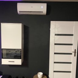 PHU ARteam Artur Rejman - Biała jednostka klimatyzacyjna Haier zamontowana na ciemnej ścianie w nowoczesnym wnętrzu, obok białych drzwi z poziomymi przeszkleniami i wiszącej szafki z ekspozycją na wino.