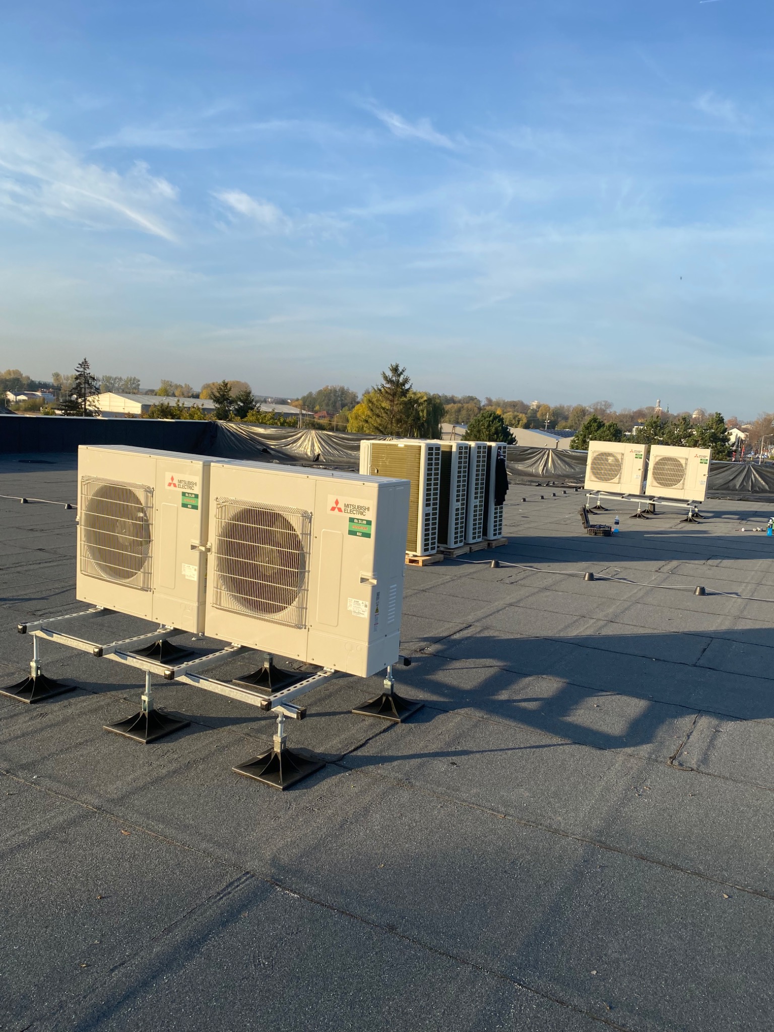 Kilka jednostek zewnętrznych klimatyzacji Mitsubishi Electric, zamontowanych na dachu budynku, widok z niskiej perspektywy, błękitne niebo w tle.