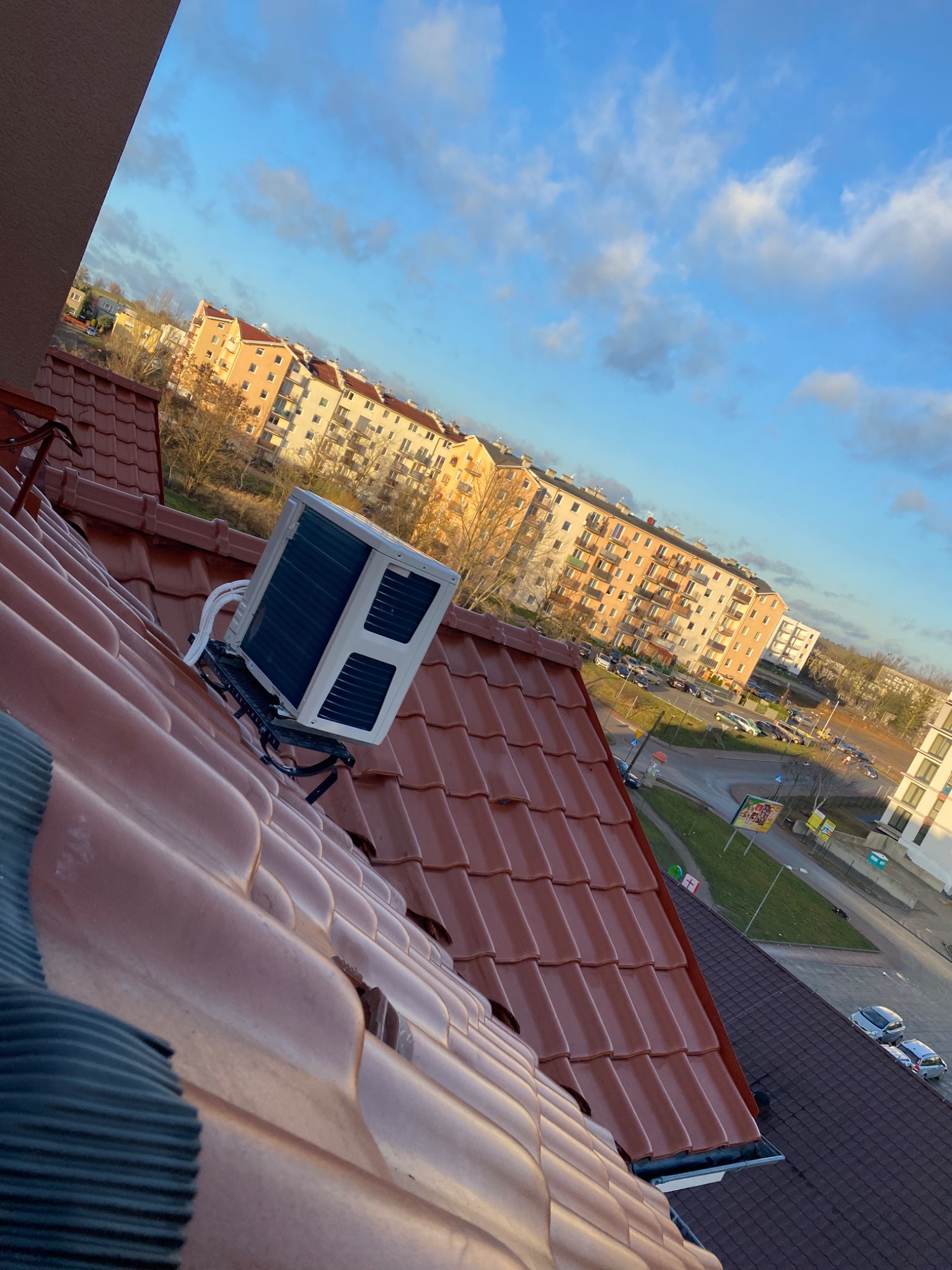Jednostka zewnętrzna klimatyzacji zamontowana na wsporniku na dachu budynku mieszkalnego, widok z góry na tle zabudowy miejskiej i błękitnego nieba.