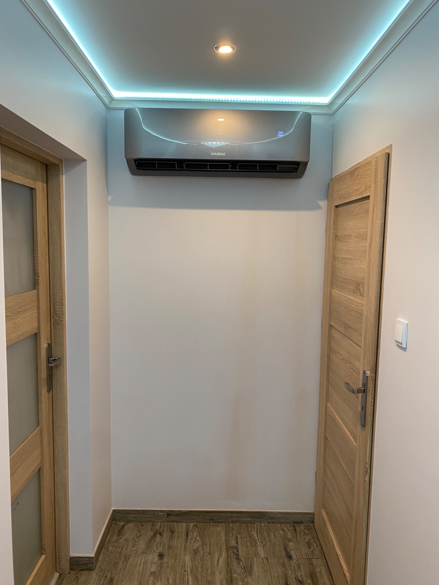 Szara klimatyzacja marki Kaisai zamontowana nad drzwiami w korytarzu z podświetlanym sufitem i drewnianymi drzwiami po obu stronach.