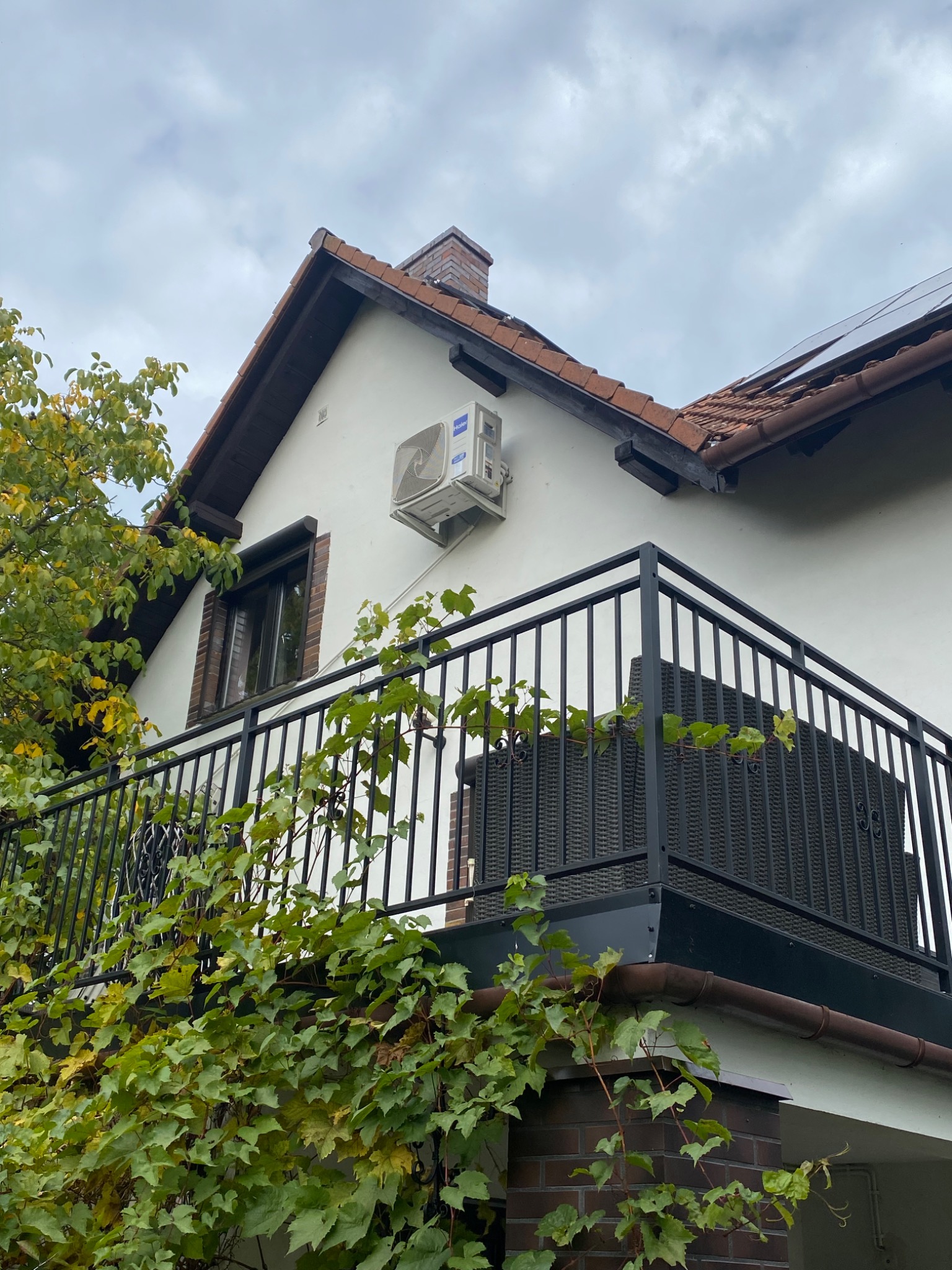 Biały klimatyzator zamontowany na ścianie domu z balkonem, widok z dołu na tle pochmurnego nieba.