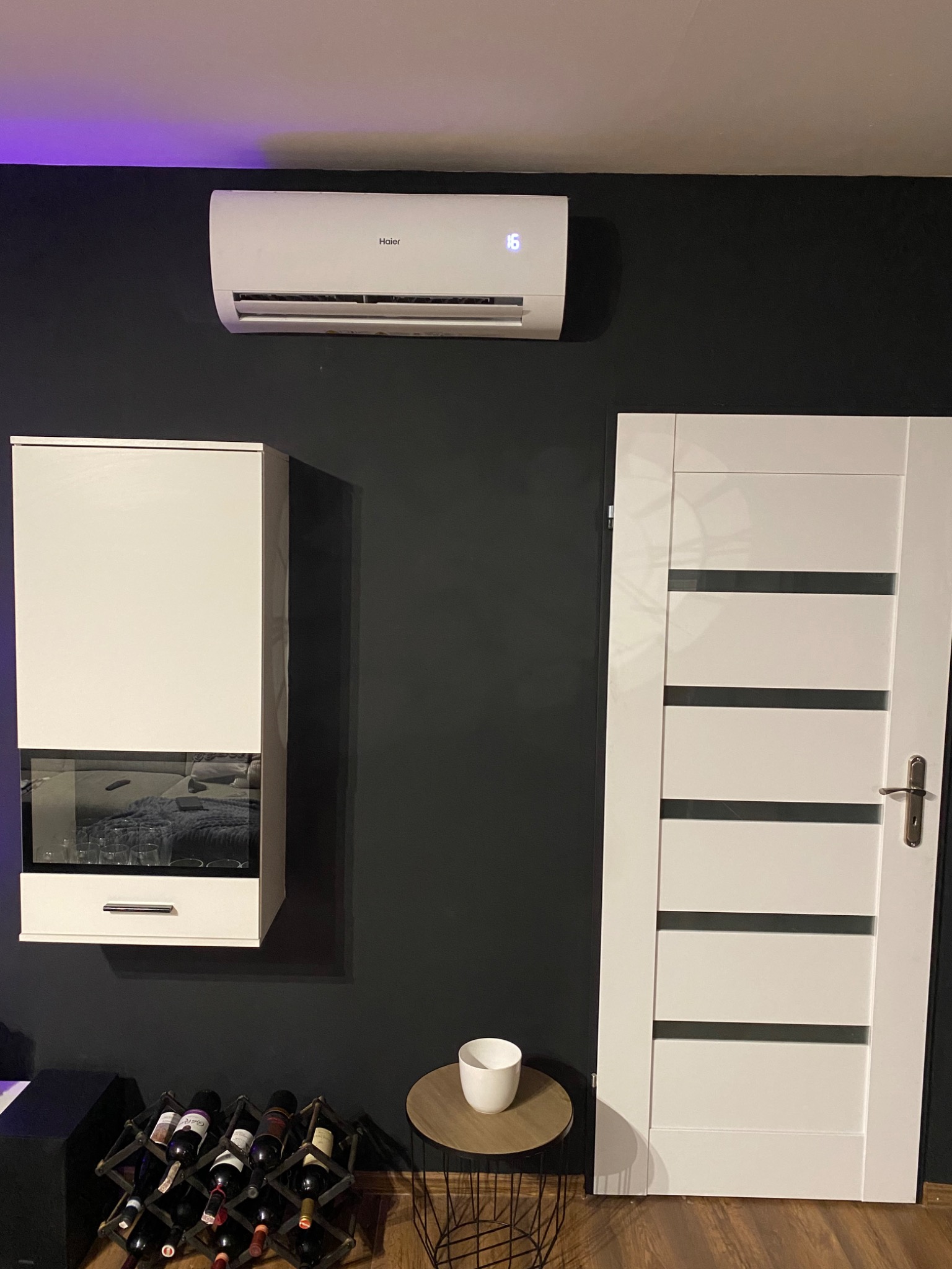 Biała jednostka klimatyzacyjna Haier zamontowana na ciemnej ścianie w nowoczesnym wnętrzu, obok białych drzwi z poziomymi przeszkleniami i wiszącej szafki z ekspozycją na wino.