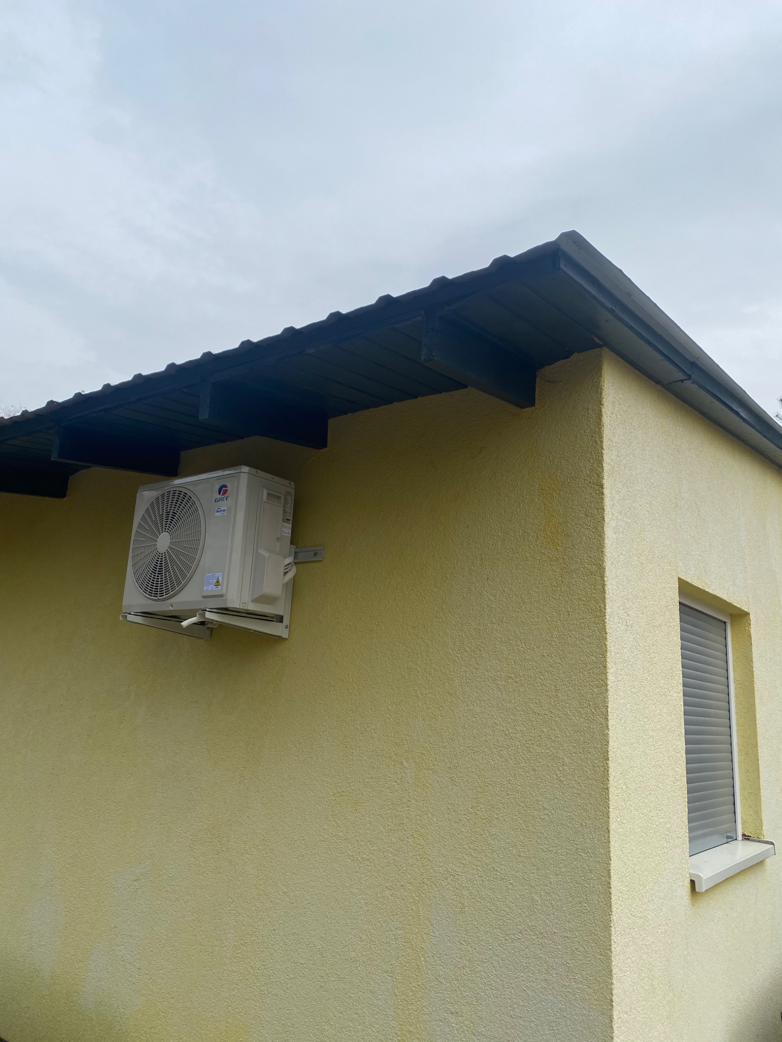 Zewnętrzna jednostka klimatyzacji marki Gree zamontowana na żółtej, teksturowanej ścianie budynku z widocznym oknem z żaluzjami i fragmentem dachu.