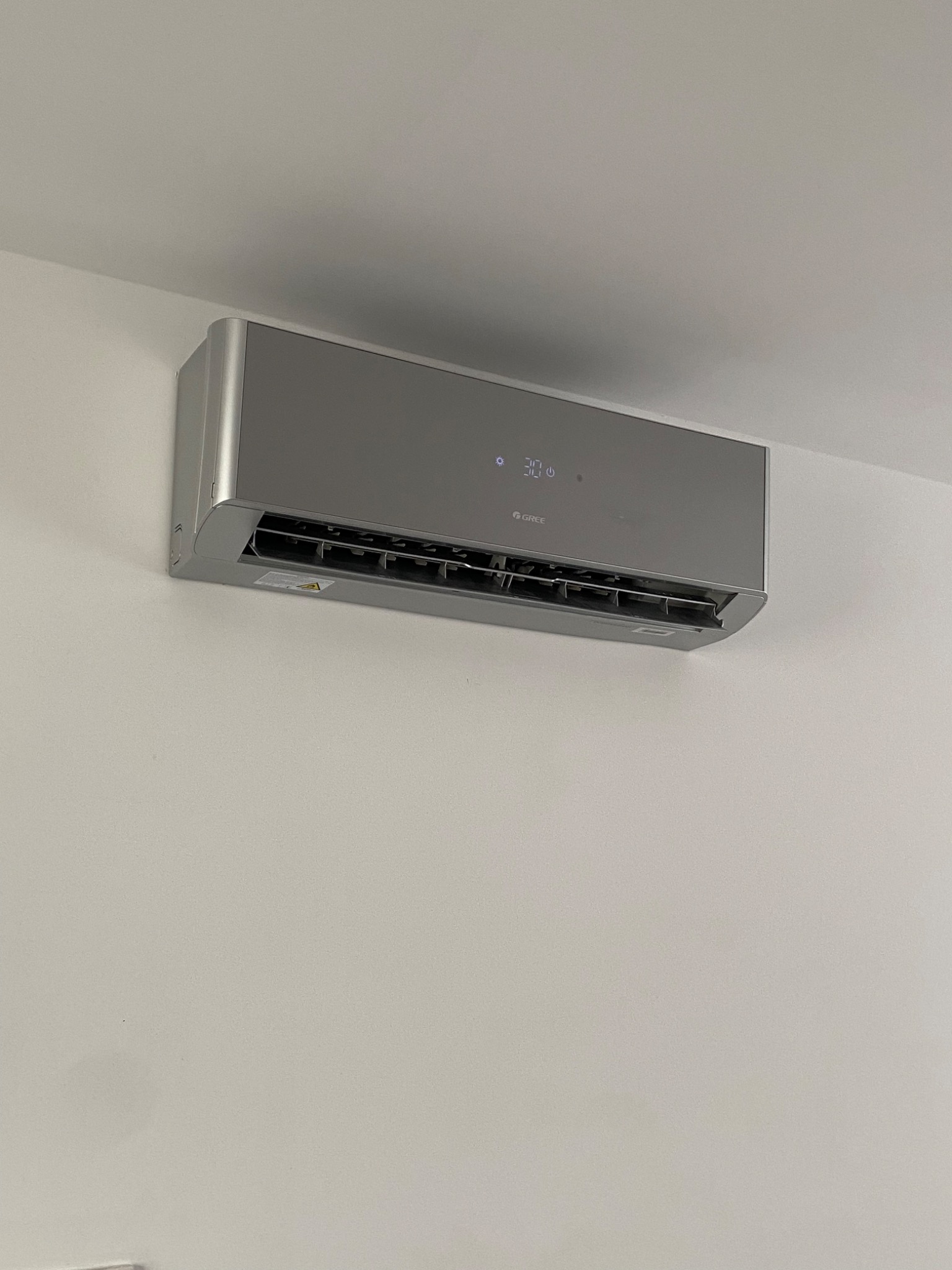 Szary klimatyzator marki Gree zamontowany na białej ścianie, wyświetlający temperaturę 30 stopni Celsjusza.