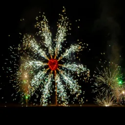 Wybuchające fajerwerki w nocy, centralny element z czerwonym środkiem i białymi iskrami tworzącymi kształt kwiatu, po prawej stronie mniejsze, złote fontanny iskier.