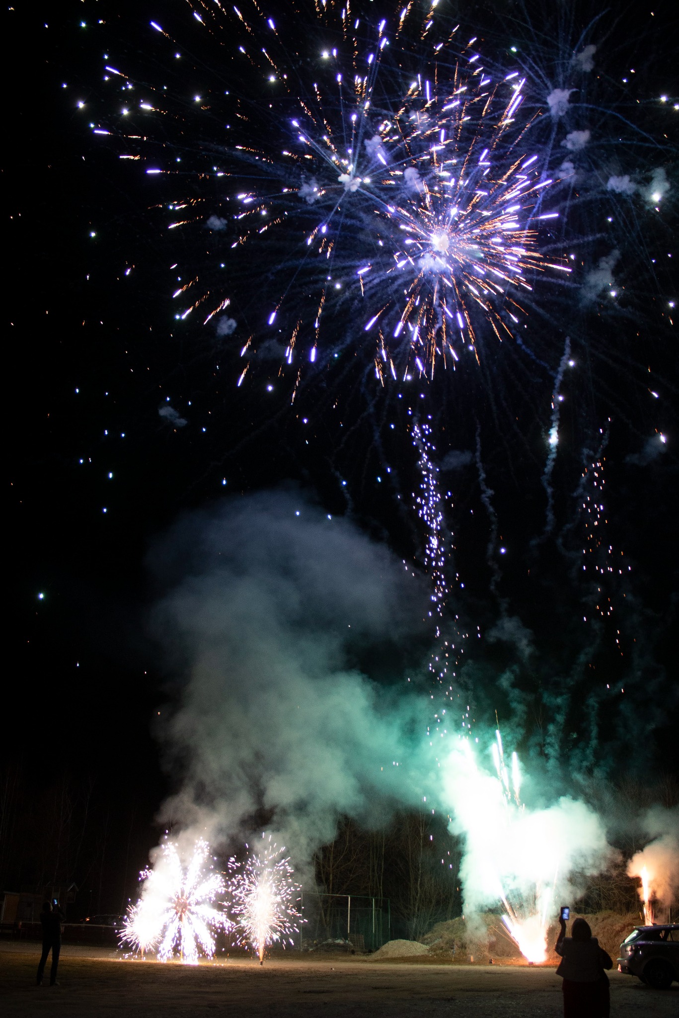 Nocne niebo rozświetlone eksplozją wielobarwnych fajerwerków; w dolnej części kadru sylwetki osób obserwujących widowisko i nagrywających je telefonami.