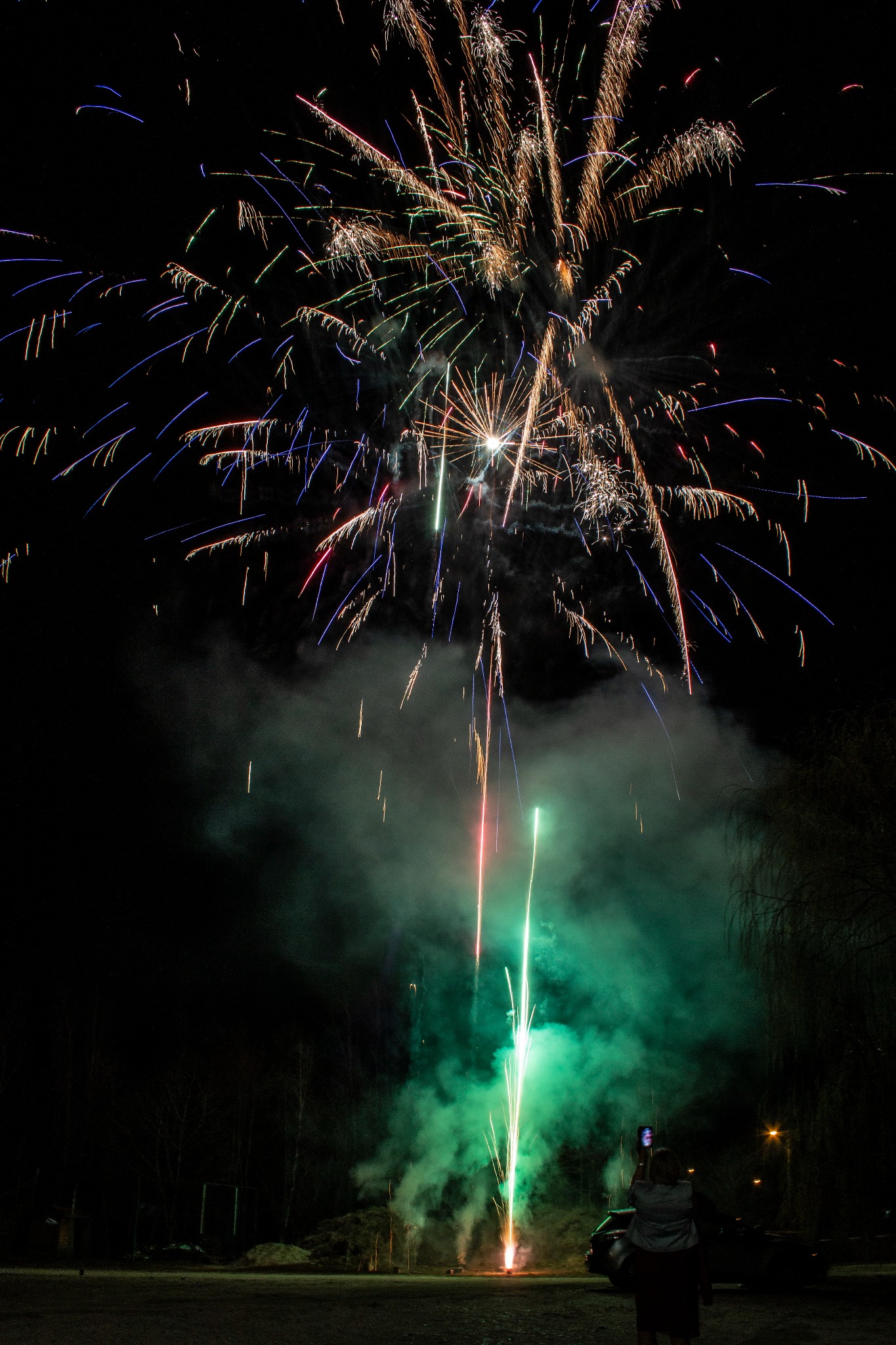 Nocne niebo rozświetlone eksplozją kolorowych fajerwerków, z zielonym dymem unoszącym się w dolnej części kadru, a w tle sylwetka osoby robiącej zdjęcie telefonem.