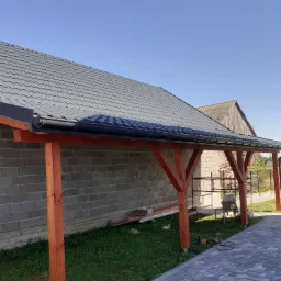 Zadaszenie tarasu z szarą blachodach&oacute;wką, drewnianą konstrukcją wsporczą i czarnymi rynnami, widoczne z boku na tle murowanego budynku i niebieskiego nieba.