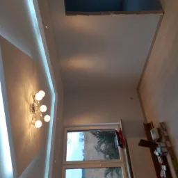 Wnętrze pokoju z podwieszanym sufitem z oświetleniem LED, widoczne także lampy sufitowe w stylu retro, okno z widokiem na zewnątrz i fragment podłogi z panelami oraz kartony w rogu pokoju.