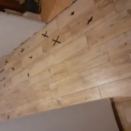 Ściana w trakcie wykańczania z ułożonymi płytkami imitującymi drewno, z krzyżykami dystansowymi, widok na fragment sufitu i drzwi.