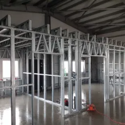 Konstrukcja ścian działowych z profili stalowych w surowym stanie, widoczne narzędzia i przewody na mokrej posadzce, metalowa konstrukcja dachu.