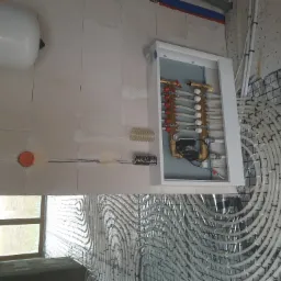 Rozdzielacz ogrzewania podłogowego w szafce, widoczne rury systemu grzewczego ułożone na izolacji podłogowej, puszka elektryczna w ścianie, zbiornik wyrównawczy.