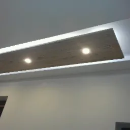 Podwieszany sufit z drewnopodobnym panelem i wbudowanym oświetleniem punktowym, otoczony listwą LED, na tle białej ściany i fragmentów okna oraz drzwi.