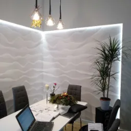 Nowoczesne biuro z falistą ścianą 3D podświetloną LED, wiszące lampy z dekoracjami, st&oacute;ł z laptopem i dekoracjami świątecznymi, roślina doniczkowa na głośniku.