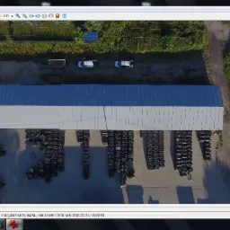 Zdjęcie z drona przedstawiające magazyn z szarym dachem, obok którego składowane są rzędy czarnych worków, a w tle widoczna droga z dwoma zaparkowanymi samochodami.