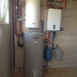 Zestaw urządzeń Ferroli: pionowy zbiornik szary, biały podgrzewacz wody i biały kocioł gazowy, połączone rurami miedzianymi i niebieskimi, z dwoma małymi zbiornikami wyrównawczymi, zamontowany...