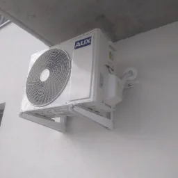 Zewnętrzna jednostka klimatyzacji marki AUX zamontowana na białej ścianie budynku, widoczne przewody i wsporniki montażowe.