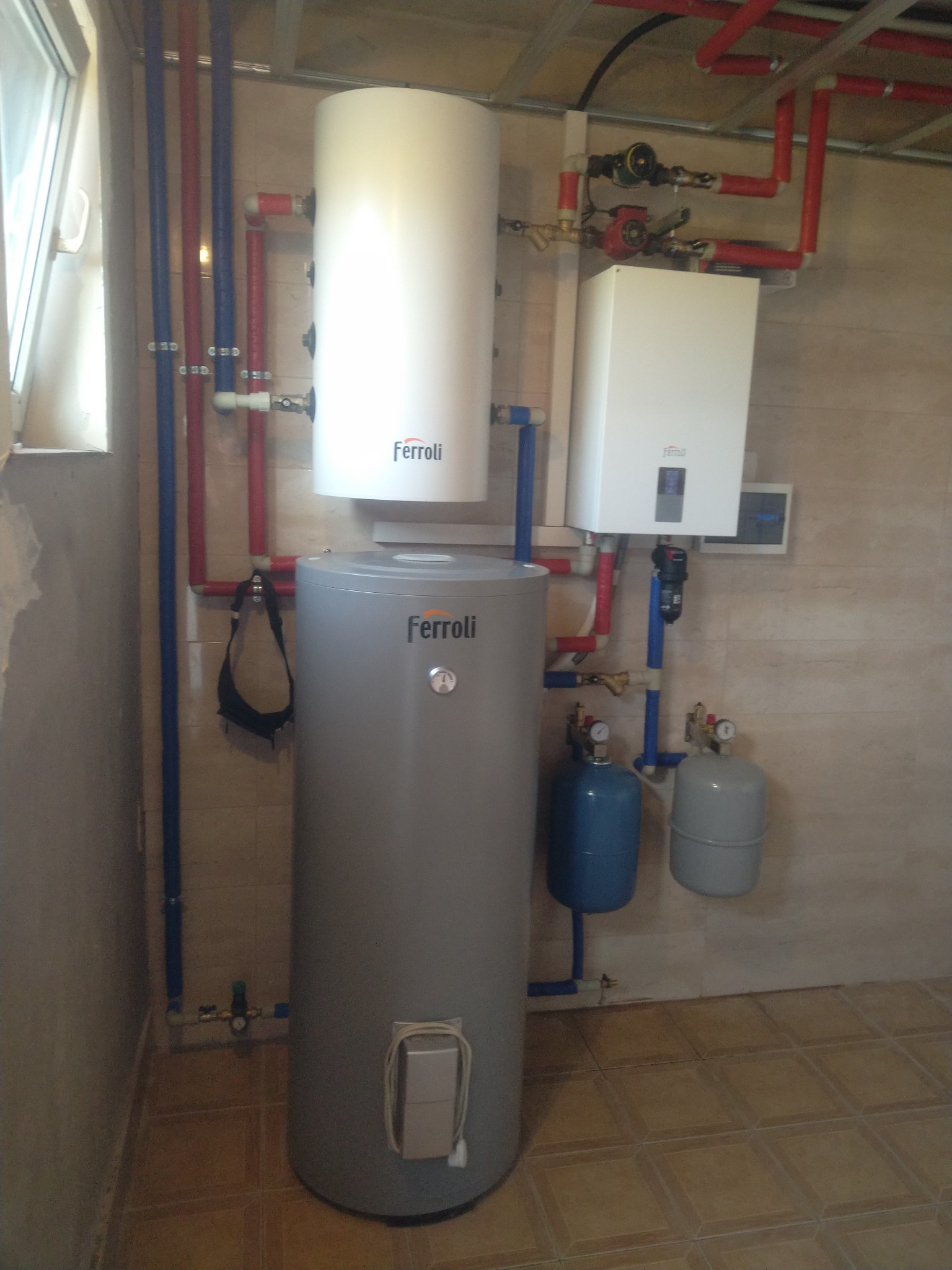 Zestaw urządzeń Ferroli: pionowy zbiornik szary, biały podgrzewacz wody i biały kocioł gazowy, połączone rurami miedzianymi i niebieskimi, z dwoma małymi zbiornikami wyrównawczymi, zamontowany...