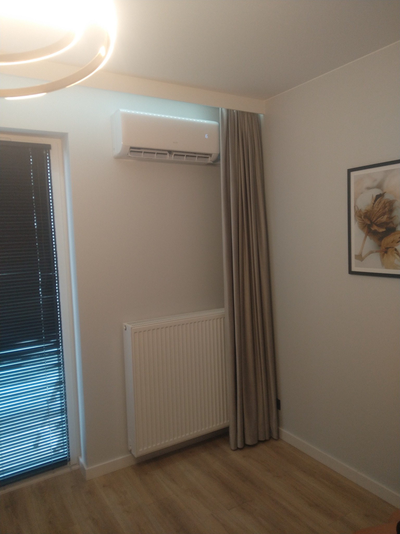 Biała klimatyzacja ścienna z oświetleniem LED nad grzejnikiem, obok zasłony i okna z żaluzjami w jasnym pokoju.