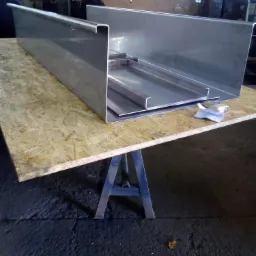 Metalowa konstrukcja kanału wentylacyjnego z blachy nierdzewnej, leżąca na drewnianym stole warsztatowym w hali produkcyjnej, z widocznymi spawami i zagięciami.