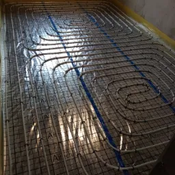 Rozłożona instalacja ogrzewania podłogowego Altech na macie izolacyjnej z folią aluminiową w trakcie montażu, widoczne białe rury grzewcze i niebieska taśma montażowa przy ścianach.