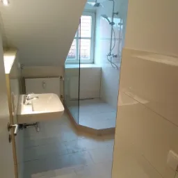 Jasna łazienka na poddaszu z kabiną prysznicową typu walk-in i umywalką nablatową, widok na okno dachowe i grzejnik.